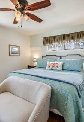 White guest bedroom with light blue and white bedding, a pink and blue floral couch and a wooden ceiling fan