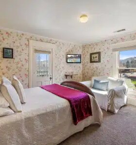 Wallpapered guest room with maroon and white bedding, a wooden closet with a mirror, and a doorway to the bathroom